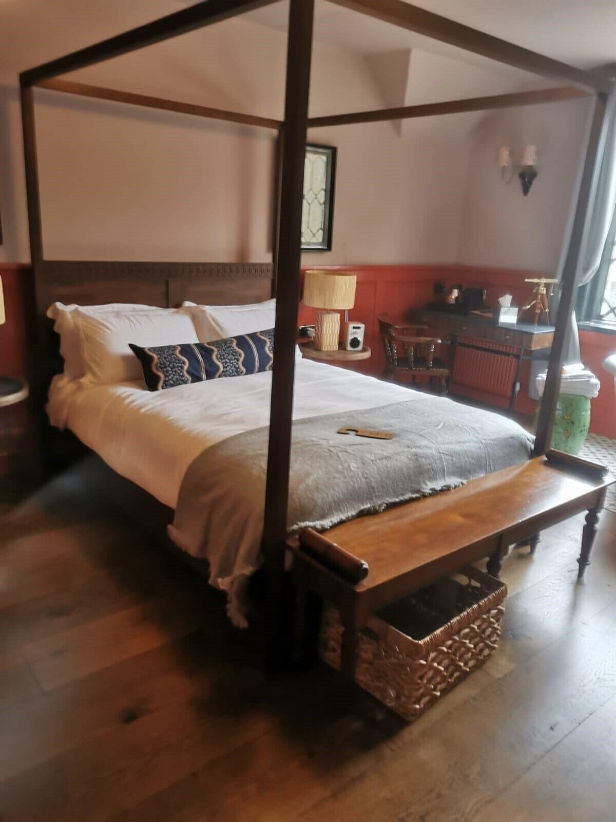 A four-poster bed with a wooden bench at the foot, a bedside table with a lamp, and a chair in a room with wooden flooring and a red wall.