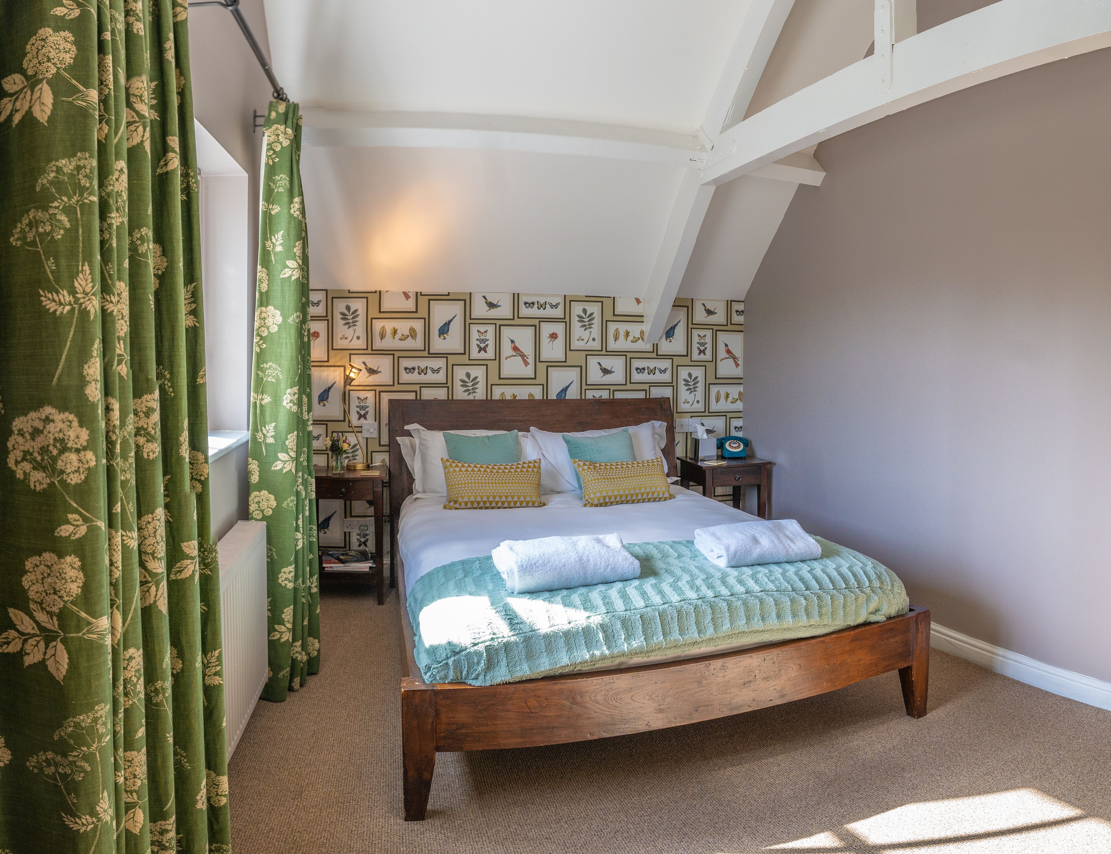 A bedroom with a wooden bed, green curtains, a patterned wall, and a nightstand with a lamp.