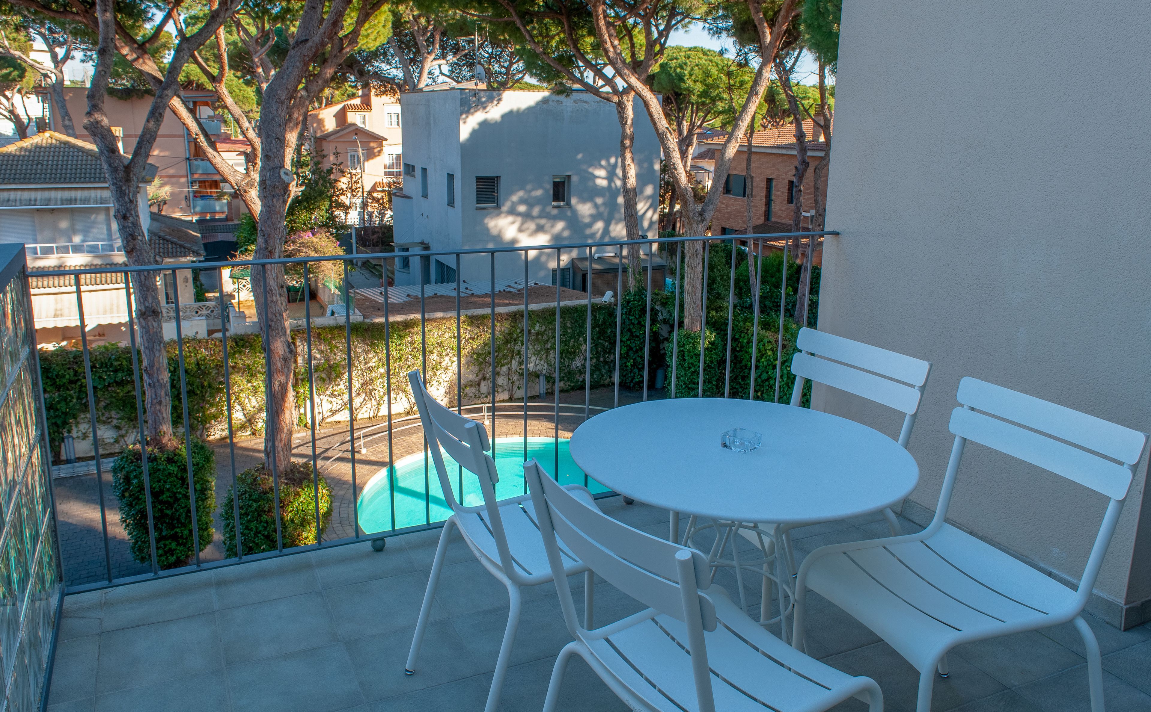 Un balcon avec une table ronde et des chaises blanches, donnant sur une piscine et des immeubles.