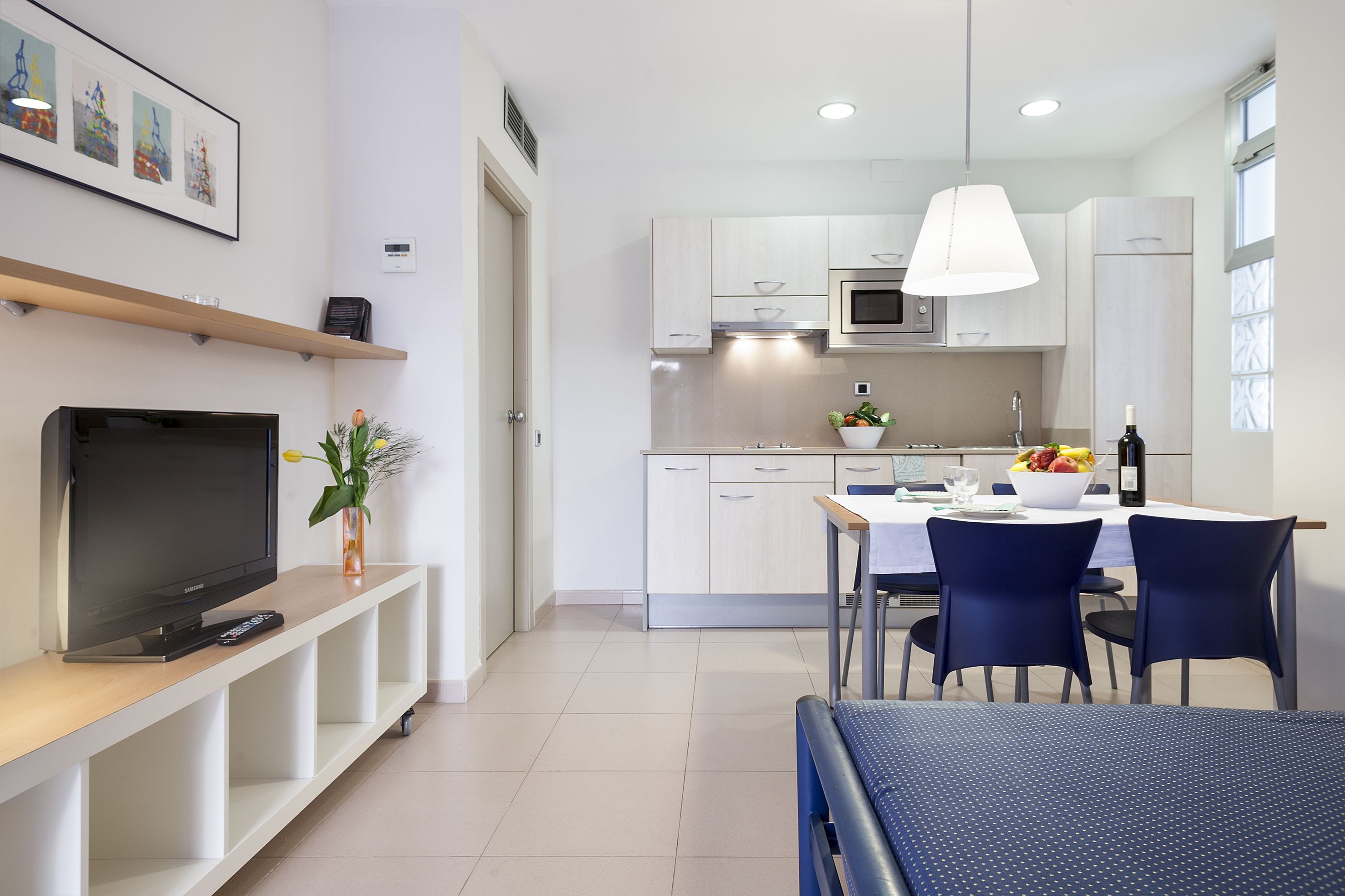 A modern kitchen with white cabinets, a dining table with chairs, a TV on a stand, and a plant.