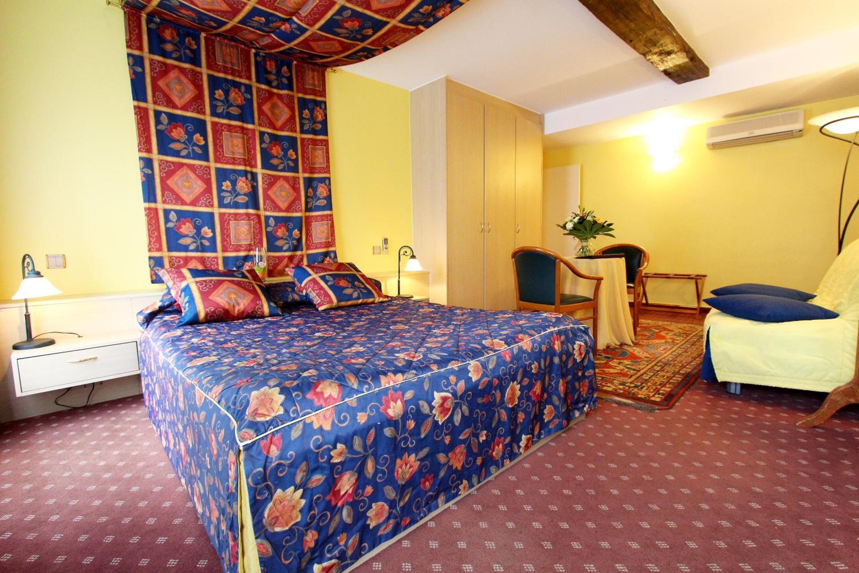 Une chambre avec une couvre-lit à motifs floraux, un bureau avec une chaise, une petite table avec une plante et un luminaire suspendu.