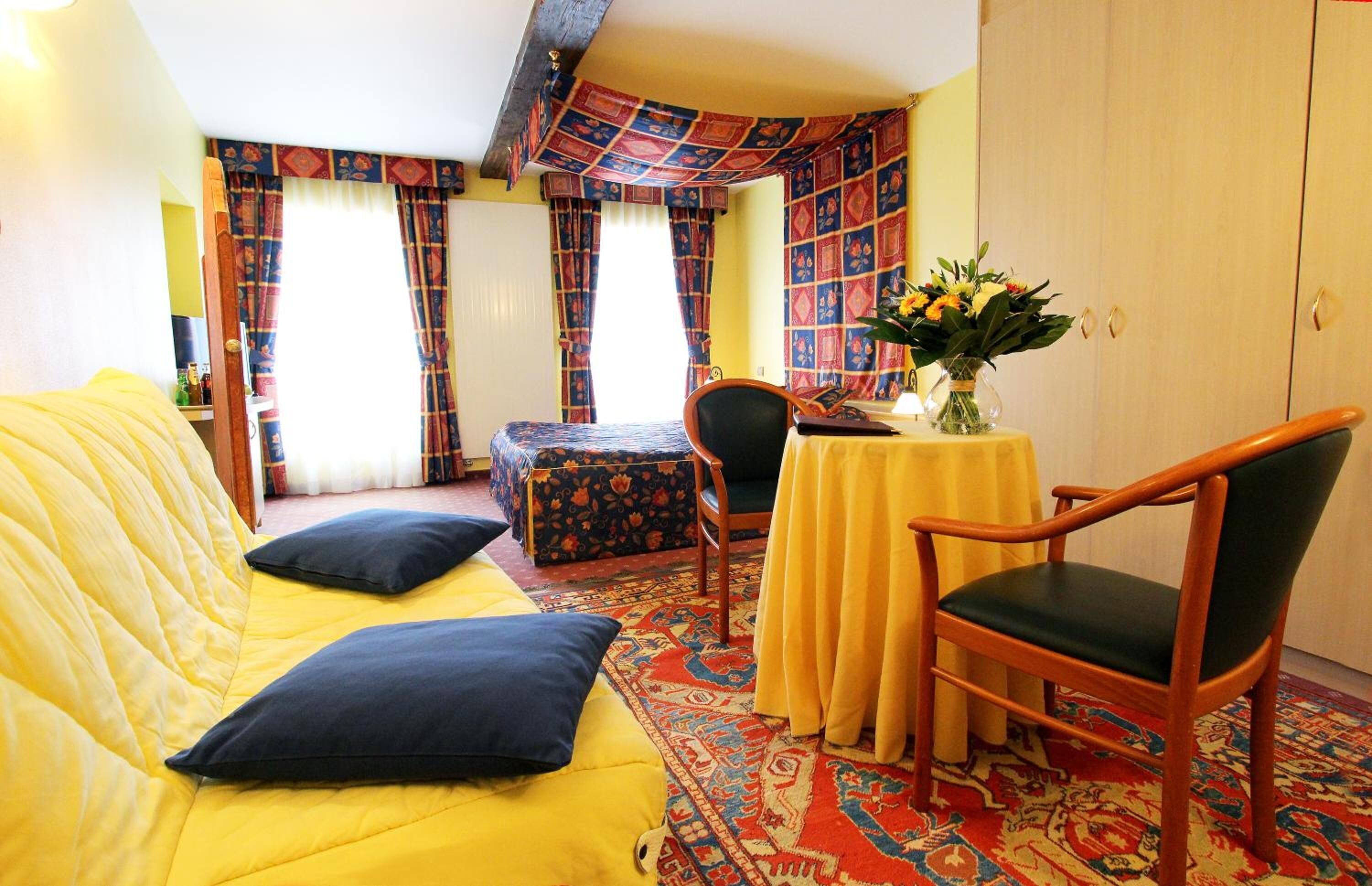 Una habitación con un sofá amarillo, una silla de madera, una mesita con un jarrón y un tapete estampado.
