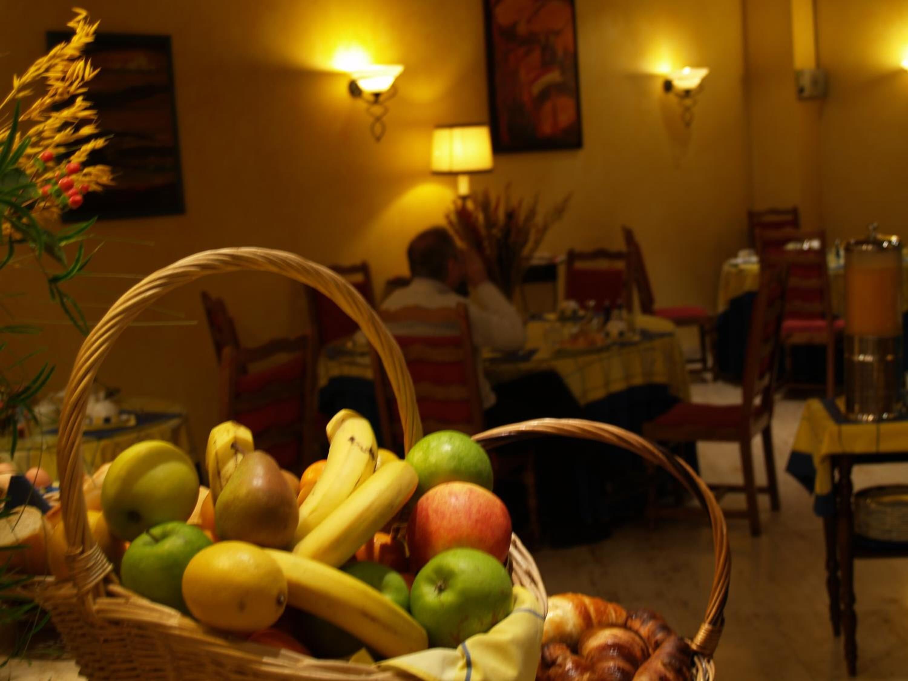 A dining area with a basket of fruits and bread on a table, a person seated at a table in the background, and wall-mounted lamps.