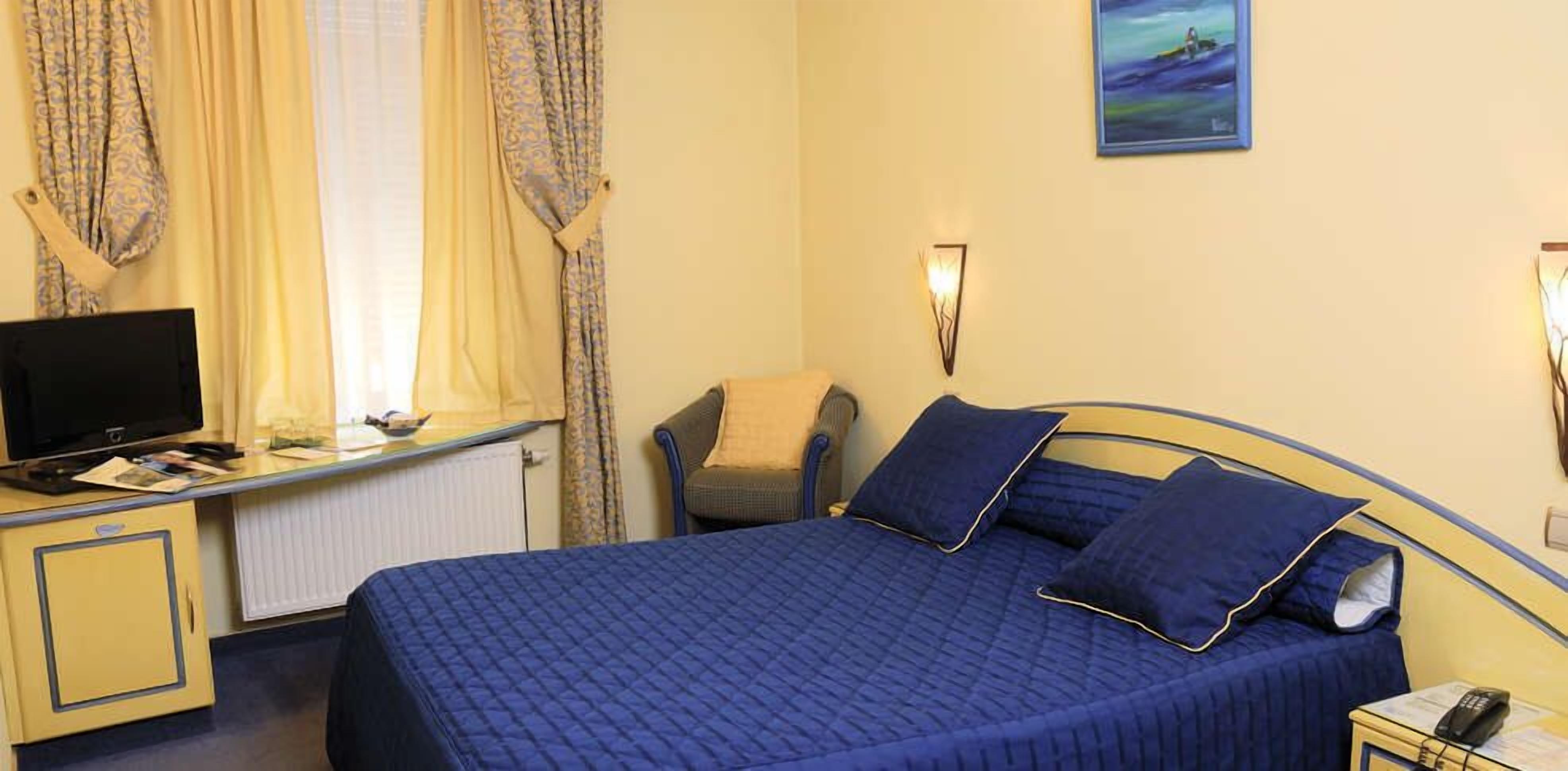 A hotel room with a bed, a desk with a computer, a chair, a window with curtains, and a painting on the wall.