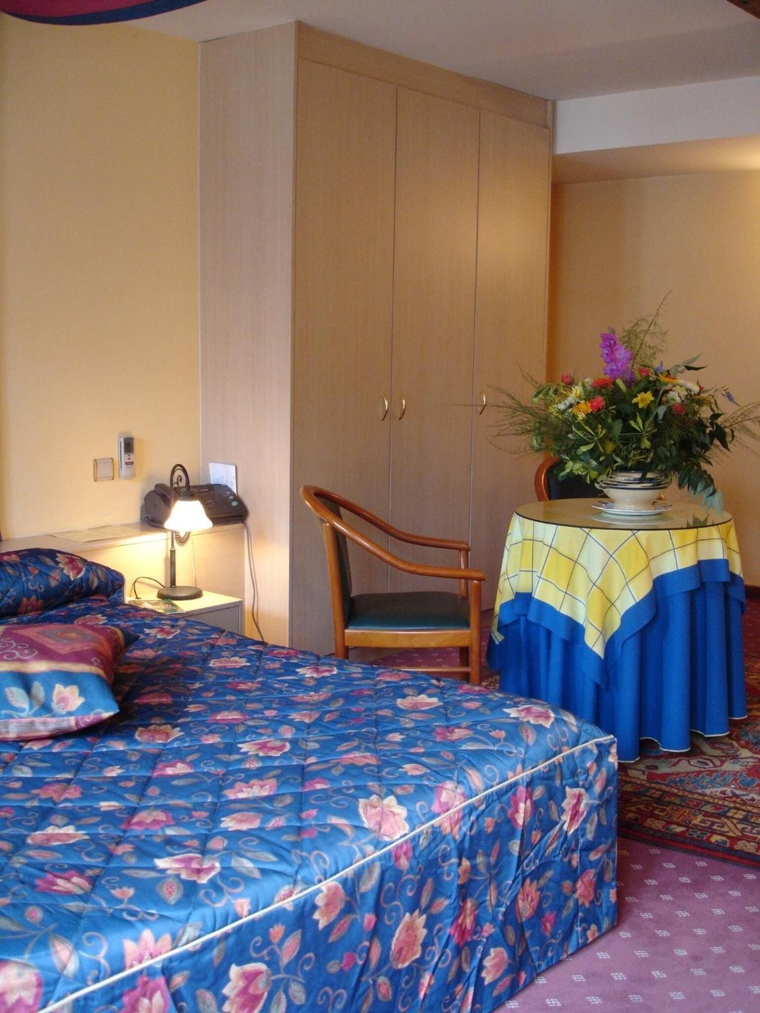 A bedroom with a bed, a chair, a table with a vase of flowers, and a wardrobe.