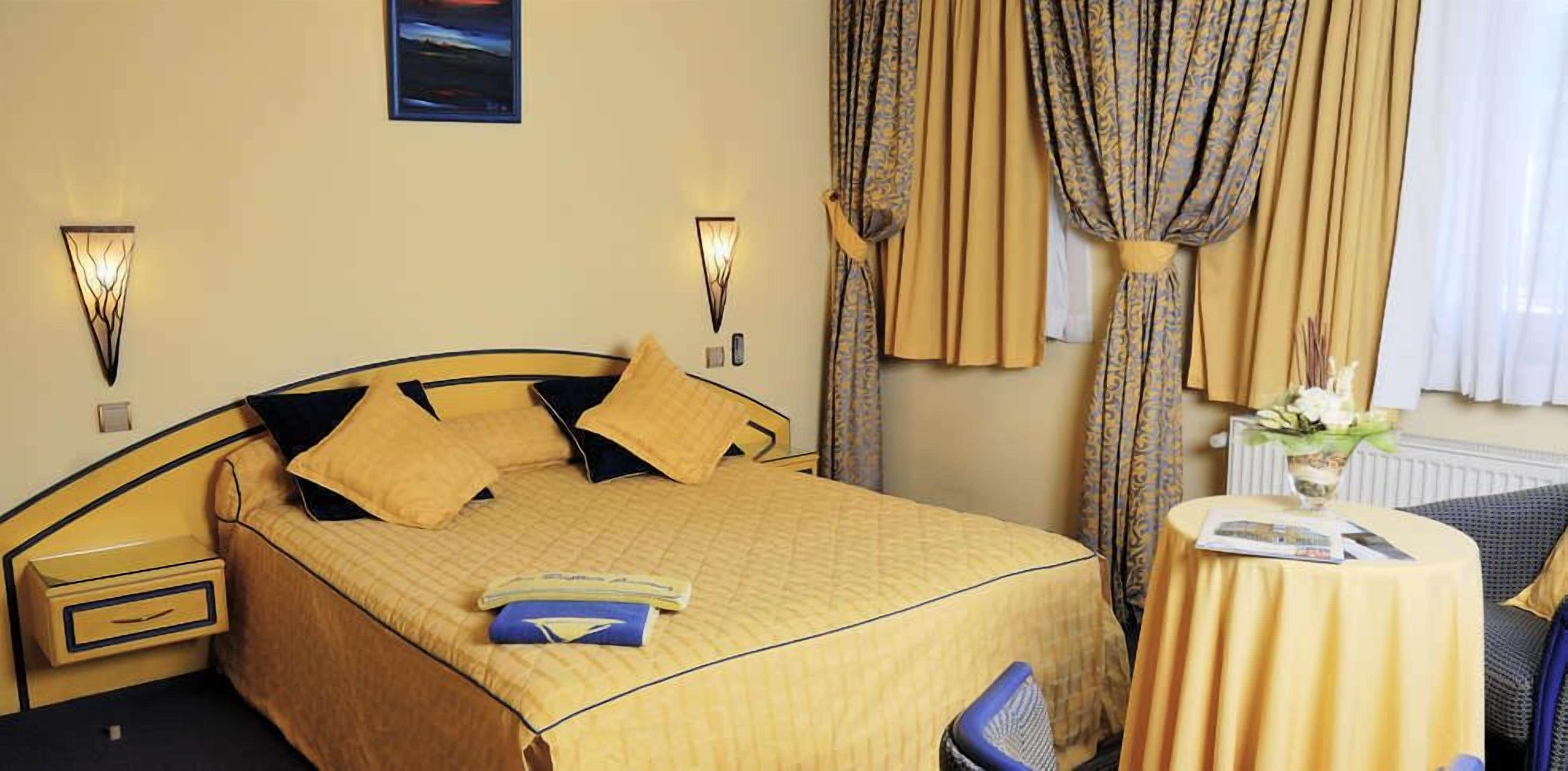 Une chambre d’hôtel comprenant un lit, une table de chevet, une commode, une table ronde avec un vase de fleurs et des rideaux.