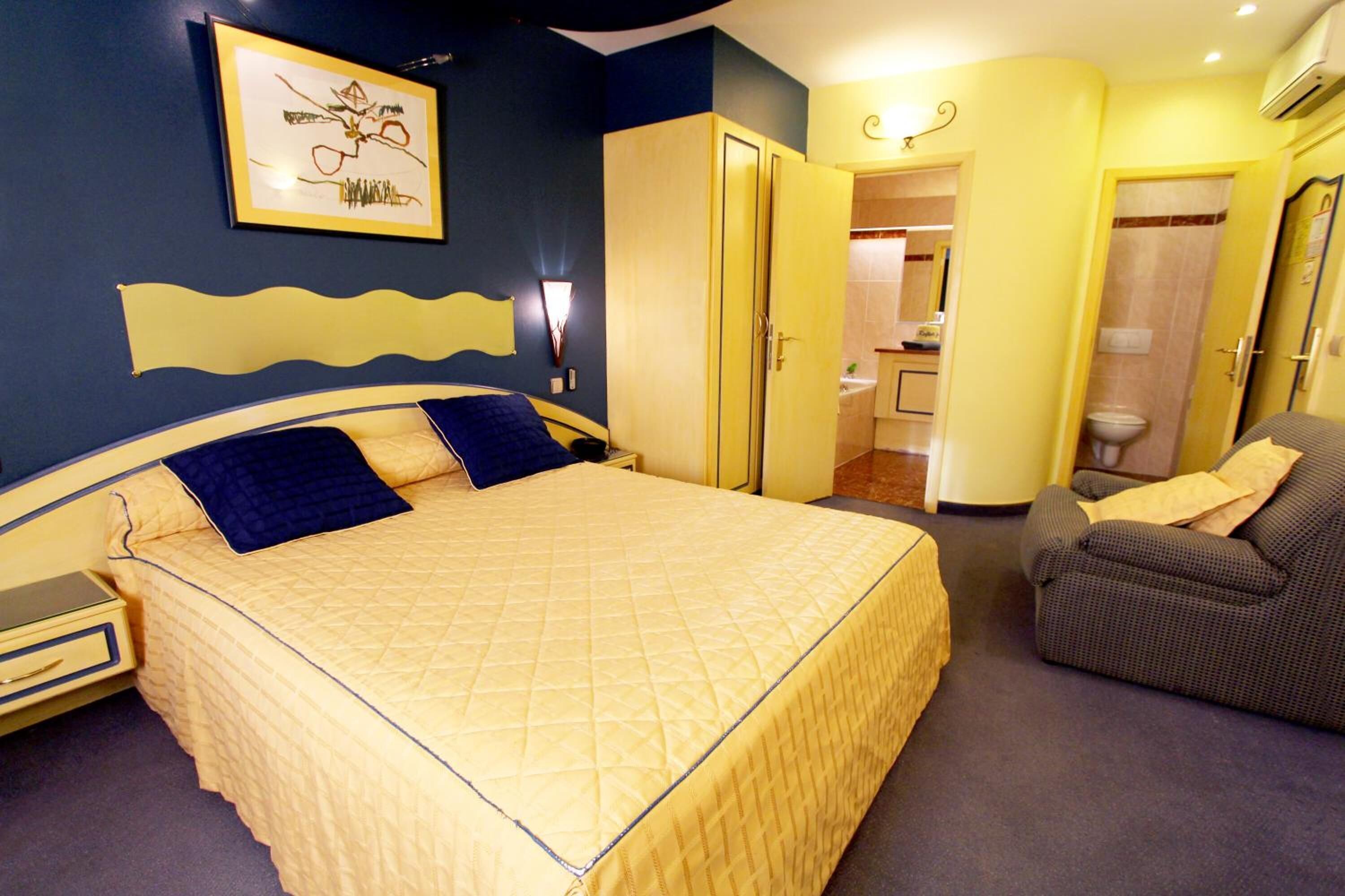Une chambre d’hôtel avec un grand lit, une chaise, une armoire et une salle de bain visible à travers une porte ouverte.