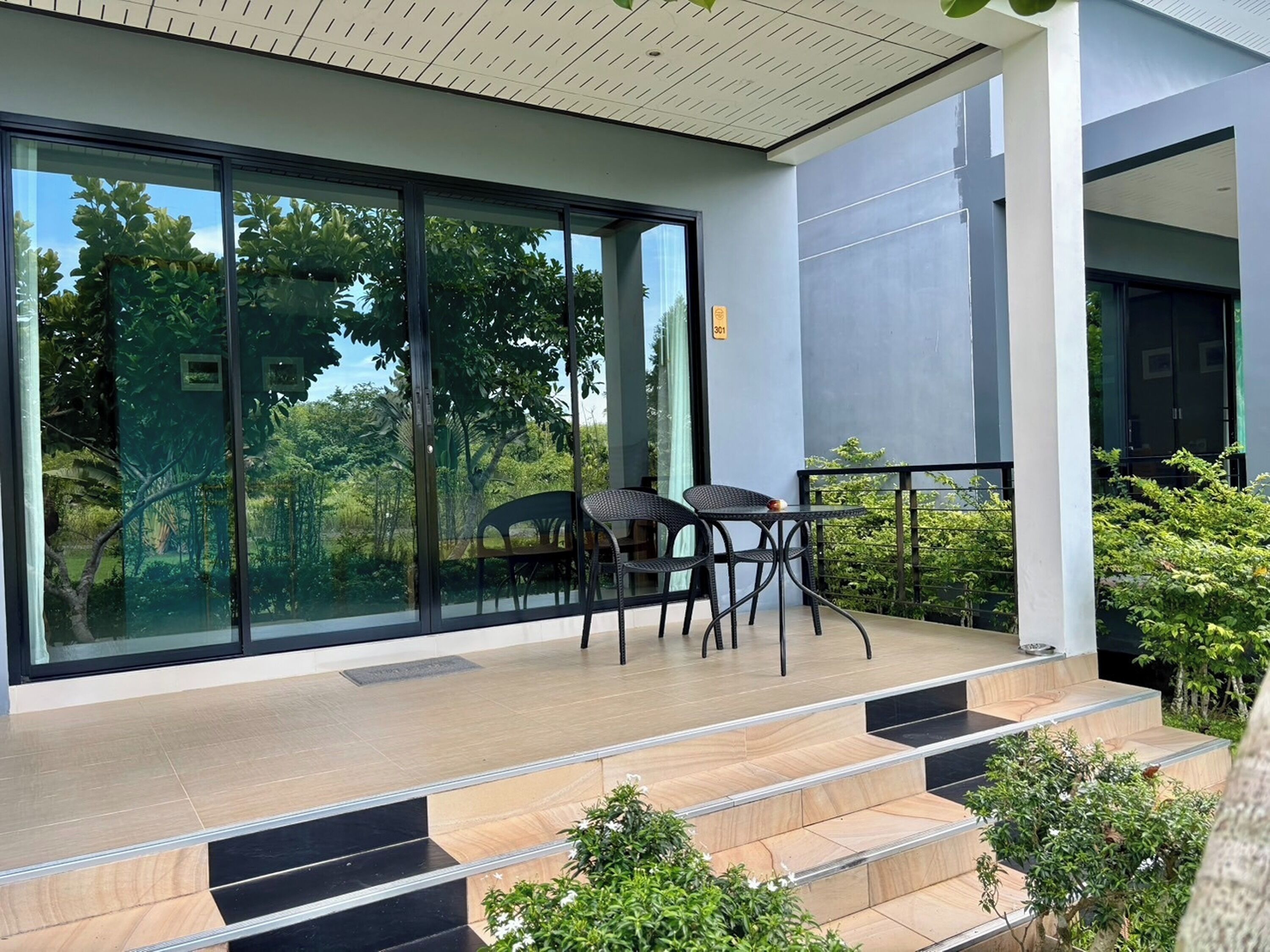 A modern outdoor balcony with a glass wall, a seating area with chairs, and a staircase leading to the entrance.