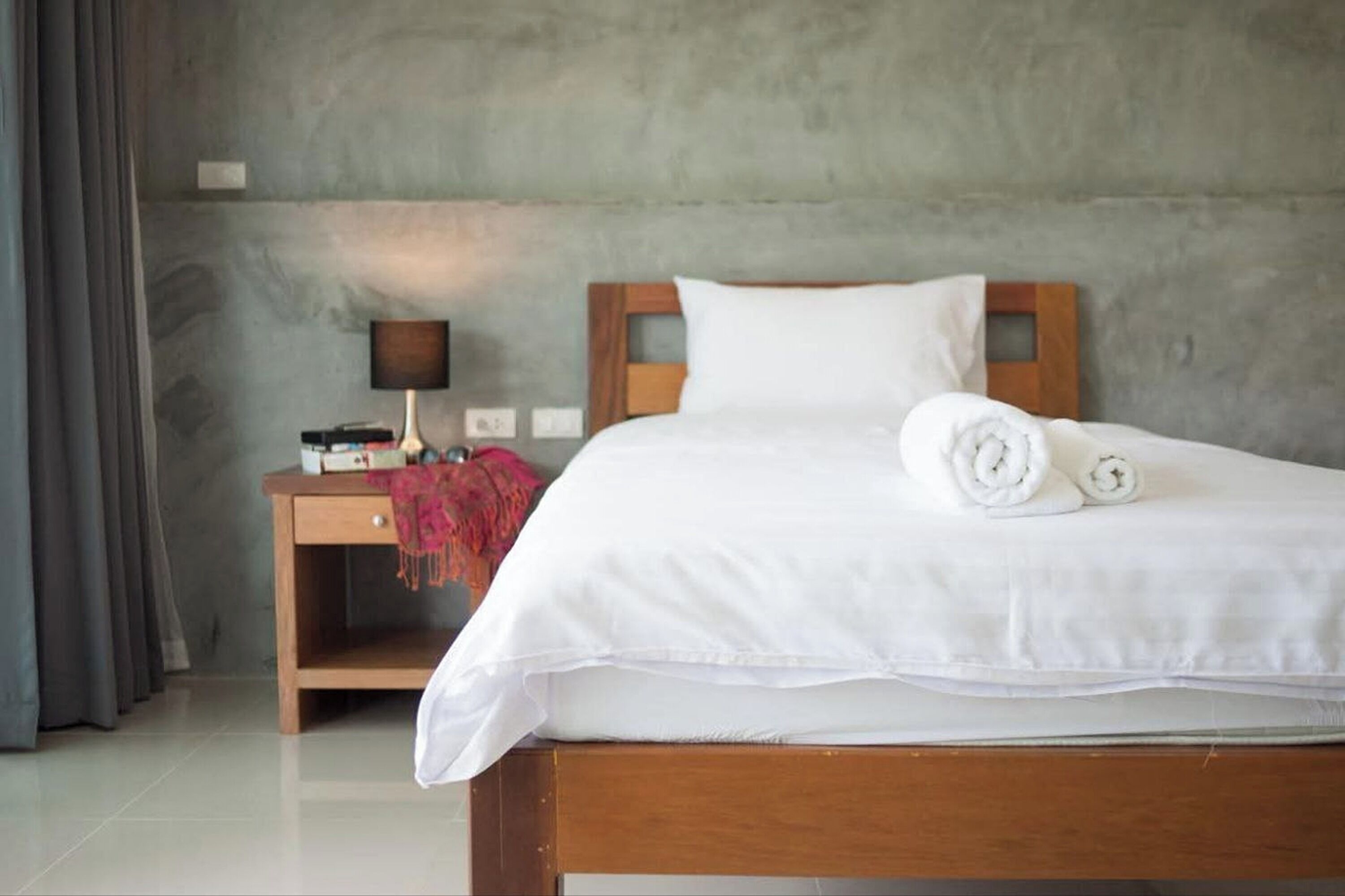 Una cama bien hecha con ropa blanca y toallas enrolladas, una mesita de noche de madera con una lámpara y una pared de concreto gris.