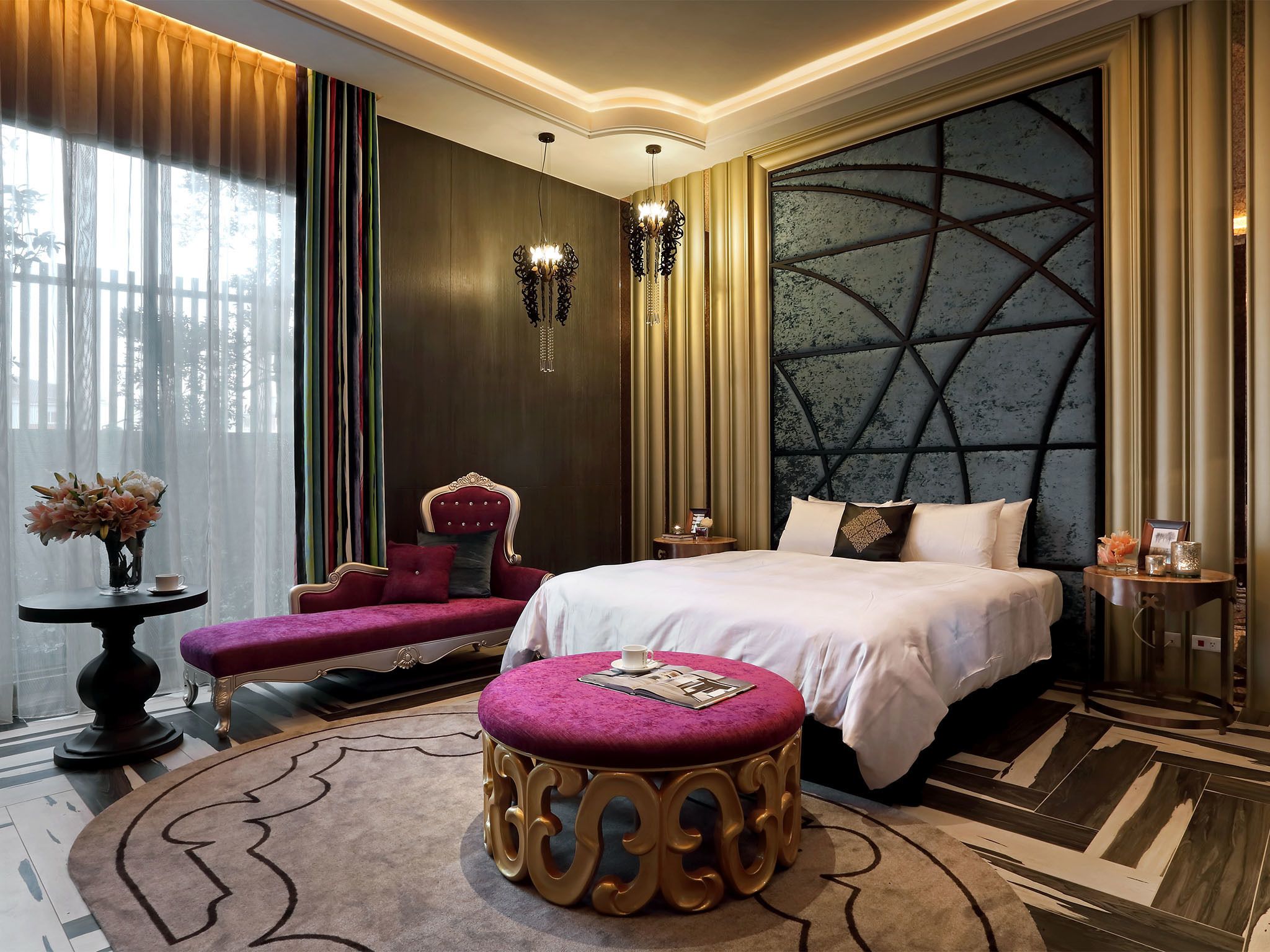 A luxurious bedroom with a large bed, a chaise lounge, a round ottoman, and a decorative wall panel.