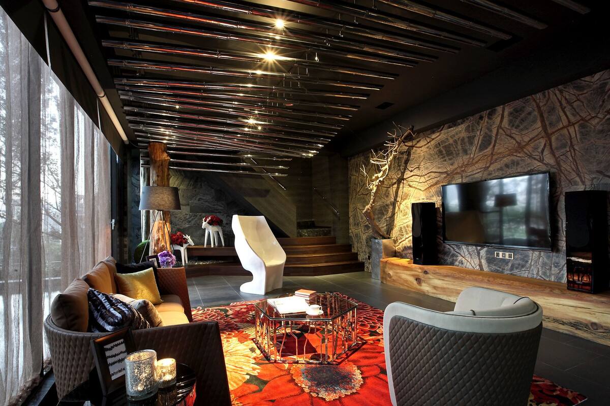 A modern living room with a sofa, coffee table, and decorative items. There is a staircase leading to another level, a TV mounted on the wall, and a large window providing natural light.
