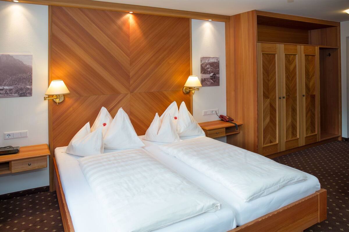 Ein Hotelzimmer mit einem großen Bett, zwei Nachttischlampen, einem hölzernen Kopfteil, einem Kleiderschrank und einem kleinen Schreibtisch mit Telefon.