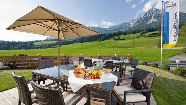 Un espace de restauration en plein air avec des meubles en osier, une table dressée avec des fruits et des aliments pour le petit-déjeuner, et une vue sur les montagnes en arrière-plan.