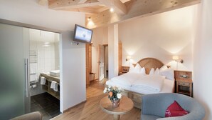 Une chambre d’hôtel moderne avec un plafond en bois, un lit recouvert de draps blancs, une petite table ronde ornée d’un vase de fleurs, un fauteuil gris et un téléviseur fixé au mur.