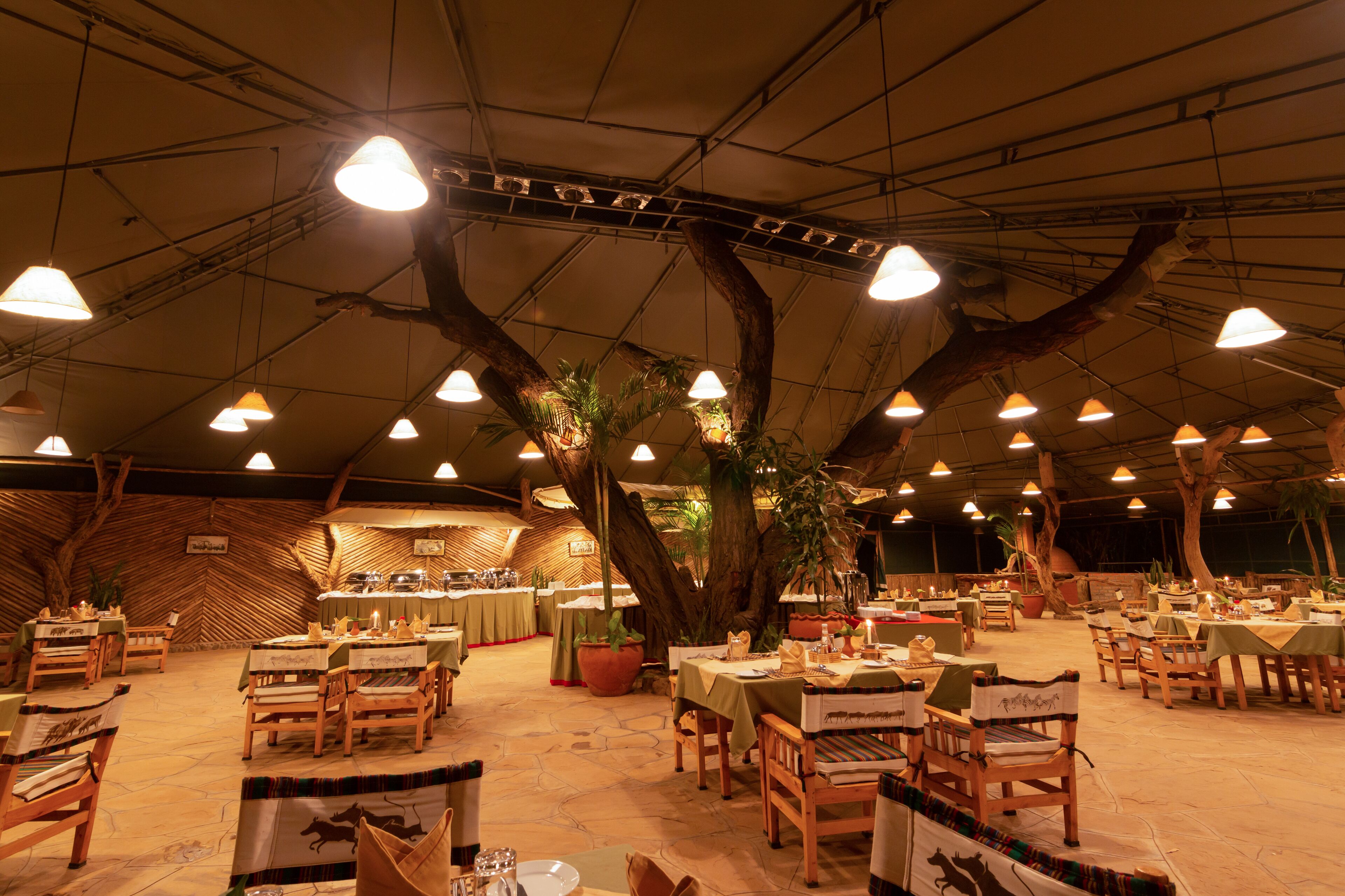 En restaurant indendørs med træborde og stole, krukker med planter og et højt loft med hængende lamper.