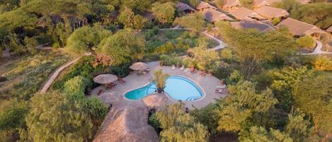 An aerial view of a resort with a central swimming pool, surrounded by thatched-roof huts and lush greenery.