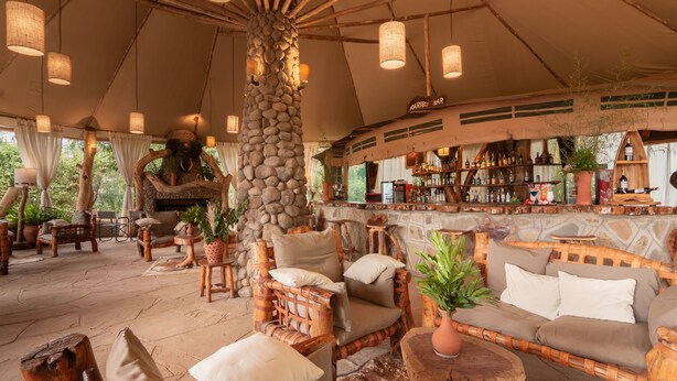 A rustic outdoor seating area with wicker furniture, a stone pillar, and a bar with various bottles. - Kibo Safari Camp (Amboseli)