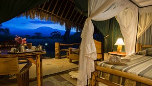 A tented accommodation with a bed, a table with a lamp, and a view of a mountain at dusk. - Kibo Safari Camp (Amboseli)
