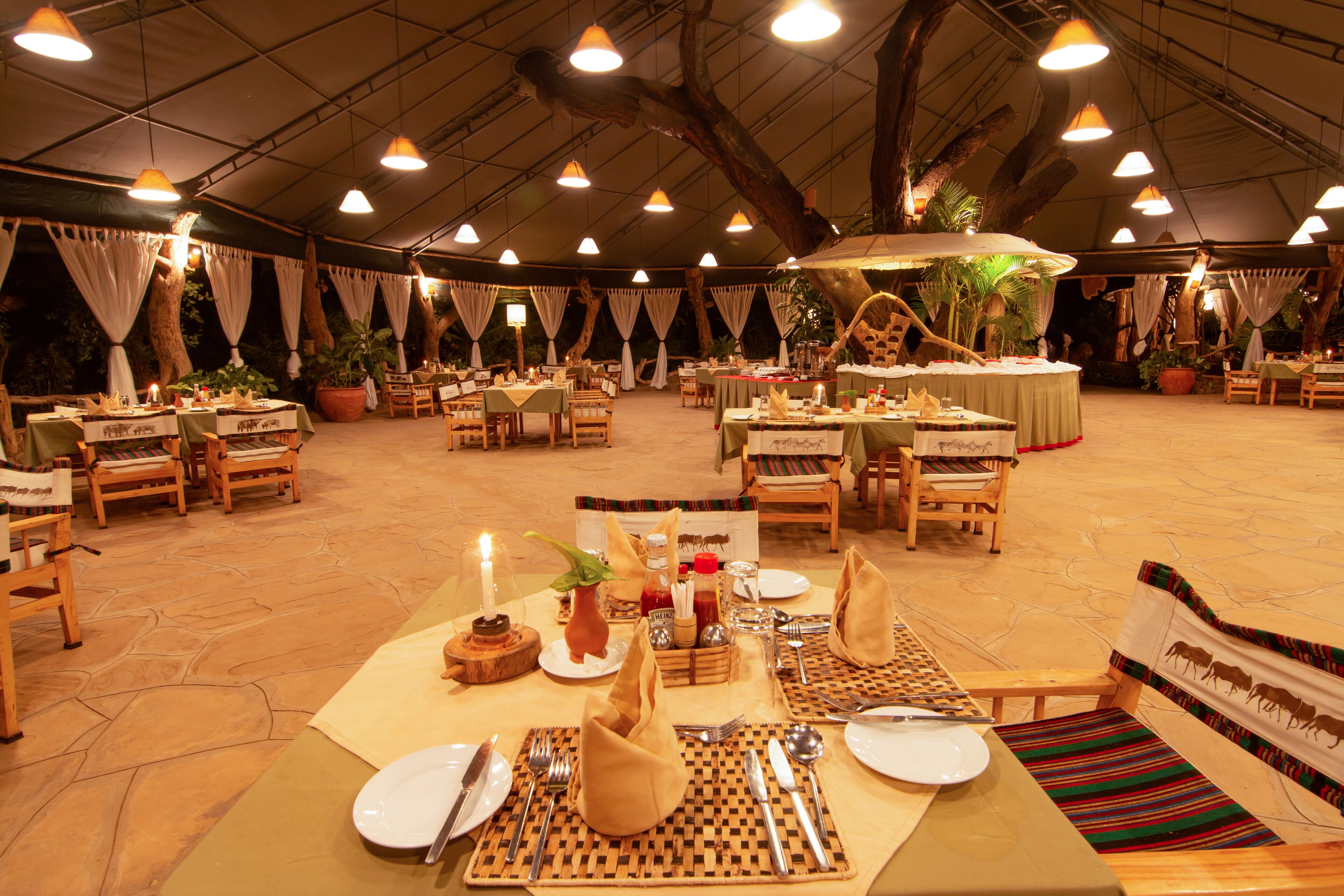 A tented dining area with tables set for multiple guests, featuring natural wood furniture and a central decorative tree.