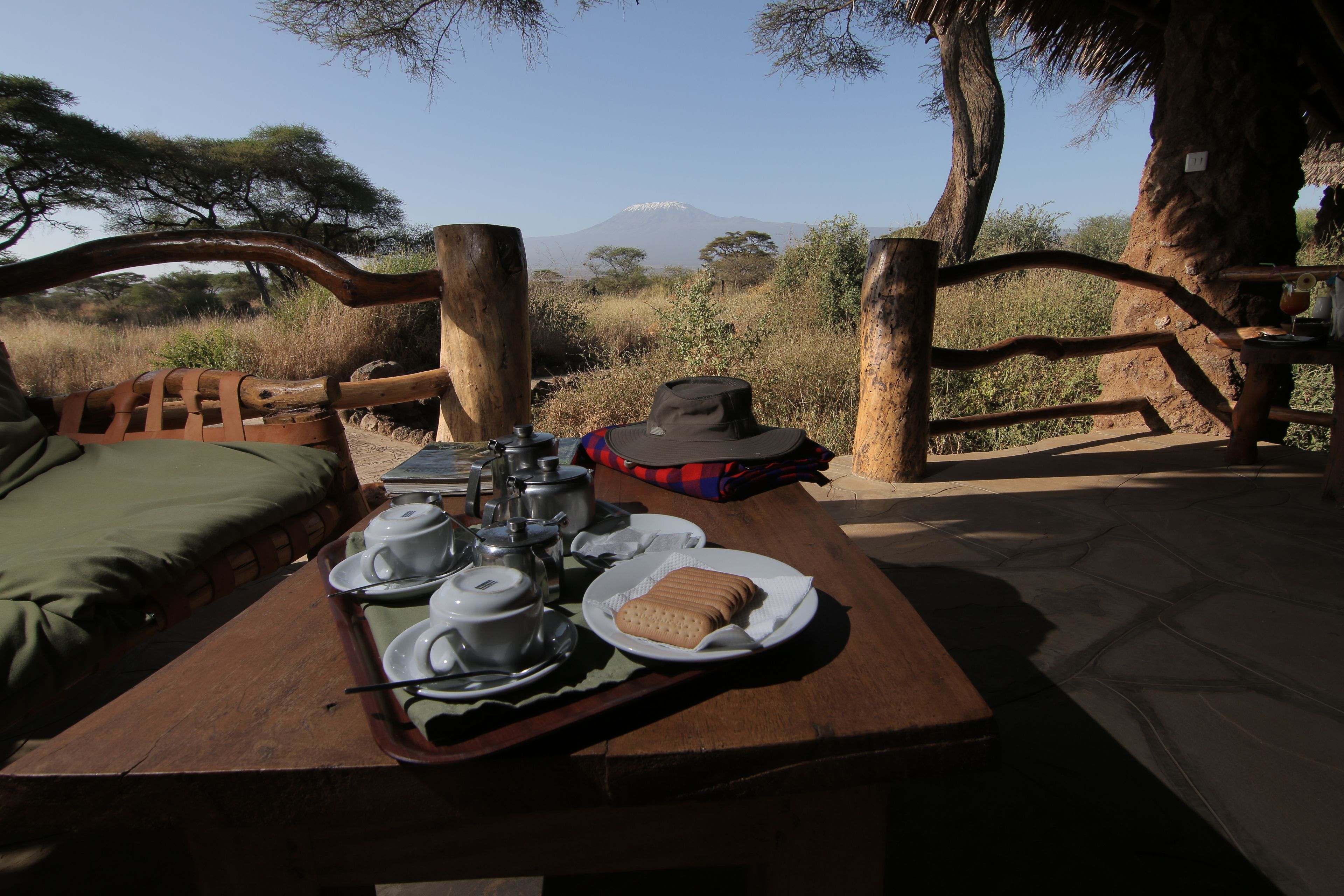 Une table de pique-nique en bois, agrémentée de services à thé, de biscuits et d’un chapeau, le tout placé devant un décor composé d’arbres et d’une montagne.