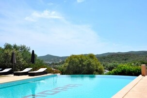 Una piscina con sillones de playa y sombrillas, rodeada de vegetación y montañas al fondo.