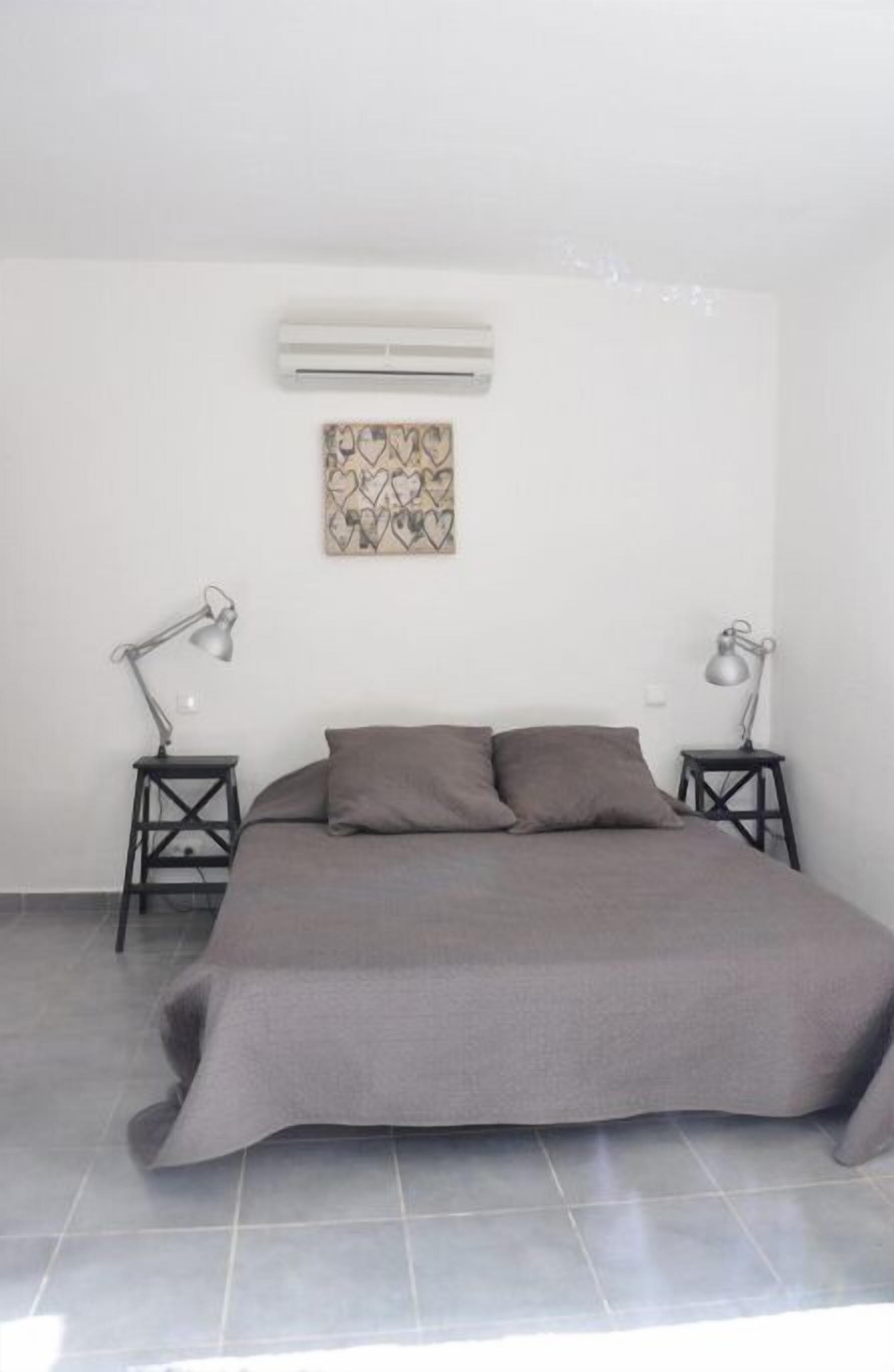 A bedroom with a bed, two bedside tables, an air conditioner, and a wall-mounted artwork.