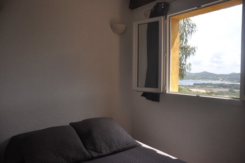 Quarto com cama, janela com vista para o mar e colinas, e luminária fixada na parede.