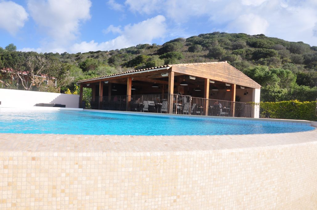 Piscina con zona de estar cubierta y un edificio al fondo.