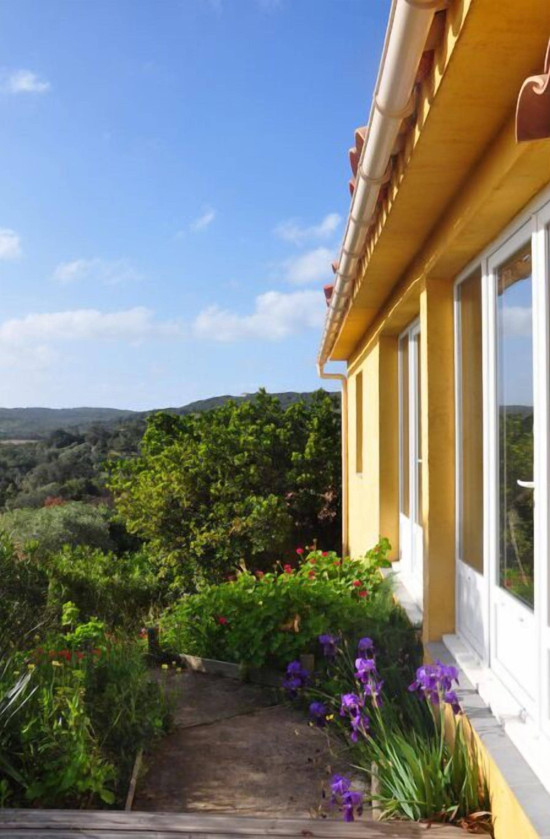 Ein gelbes Gebäude mit einem Gartenweg, der zu einem Fenster mit Blick auf eine hügelige Landschaft führt.