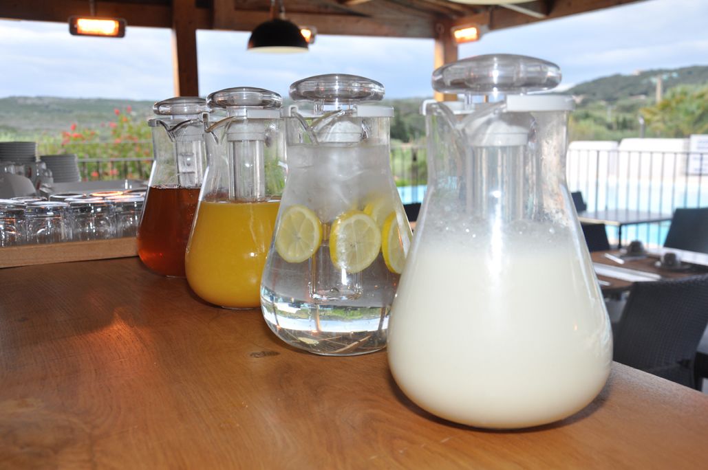 A bar area with several glass pitchers containing different beverages, a wooden table, and a view of a pool and hills in the background.