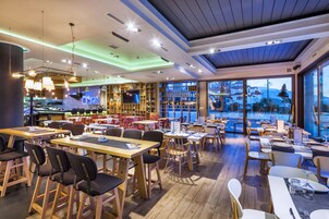 Un restaurante con mesas y sillas de madera, una zona de bar y grandes ventanales que ofrecen vistas al exterior.