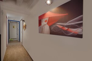 Un corridoio con pavimento in legno, una porta con cornice blu e un'opera d'arte a parete raffigurante una figura con un cappello rosso e forme astratte.