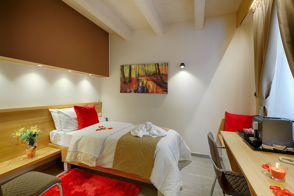 Une chambre d’hôtel avec un lit, un bureau, une chaise, un tapis rouge, une plante et un tableau au mur.