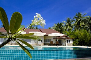 A swimming pool with tiled edges, surrounded by palm trees and a building with a tiled roof and white walls.