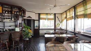 Una zona de bar con mobiliario de mimbre, ventilador de techo y grandes ventanales con cortinas.