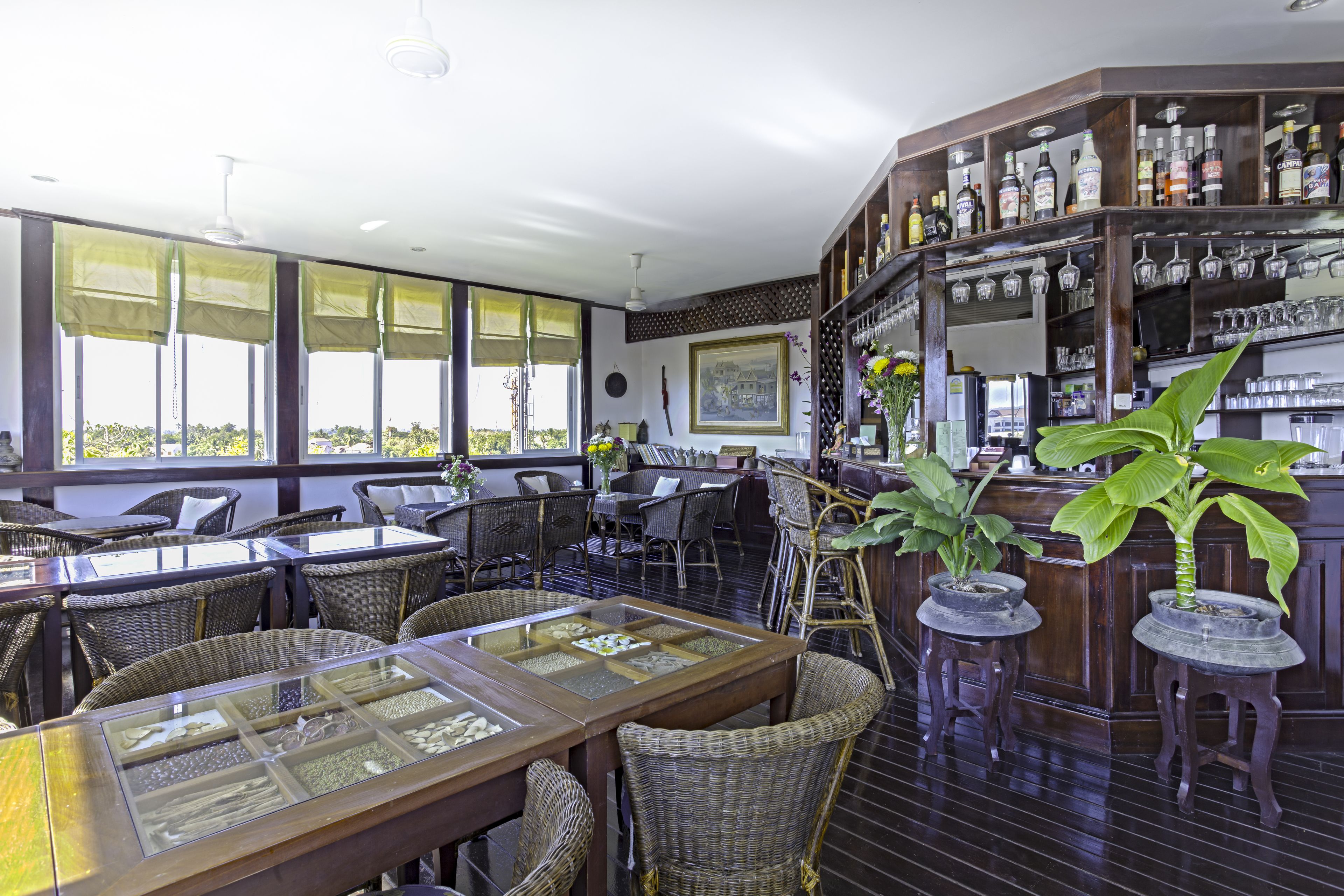 A restaurant with wicker furniture, a bar area with shelves of bottles, and large windows.