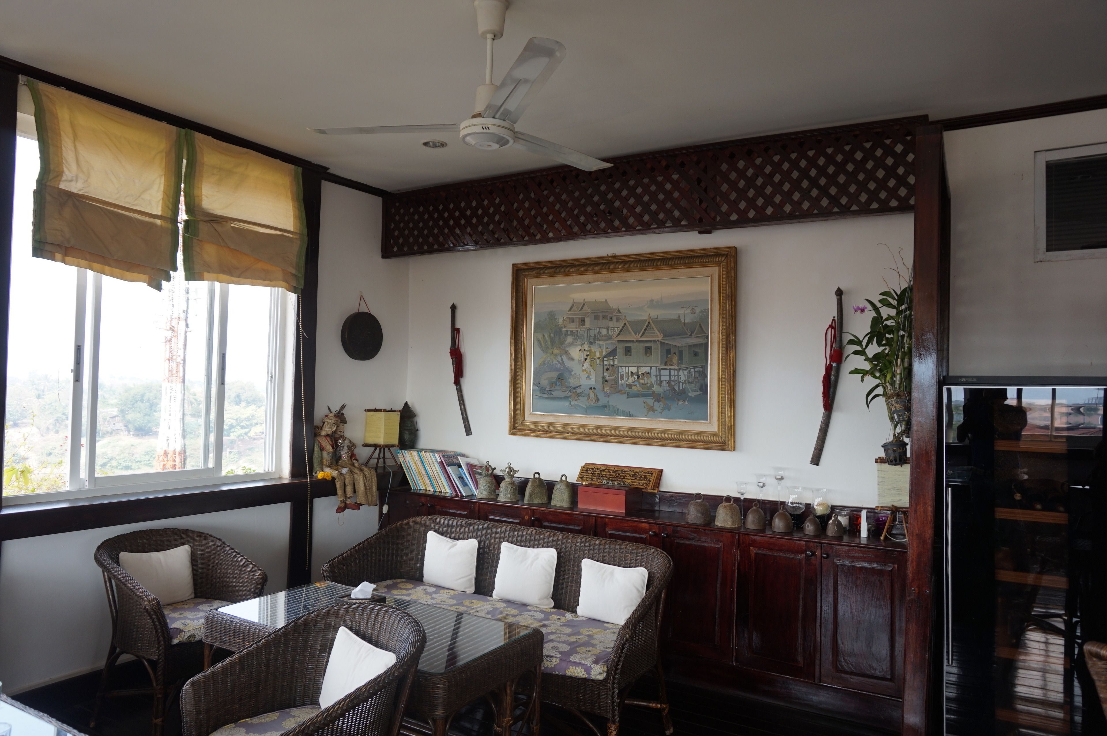Una habitación con muebles de mimbre, un ventilador de techo, un cuadro grande enmarcado y vistas por la ventana.