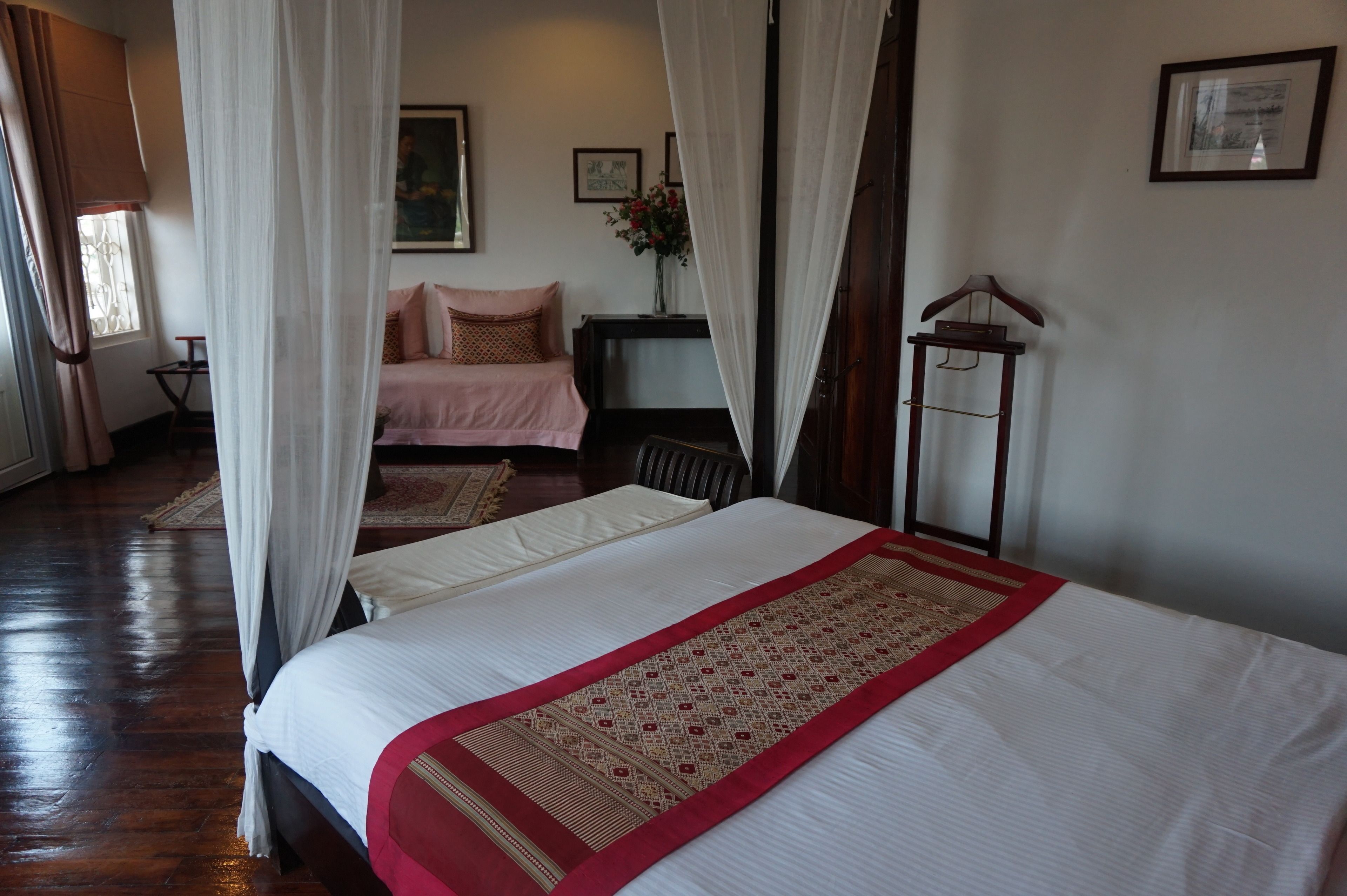 A room with two beds, one with a red and beige bed runner, and the other with a pink bedspread. There is a wooden chair and a small table with a vase of flowers.
