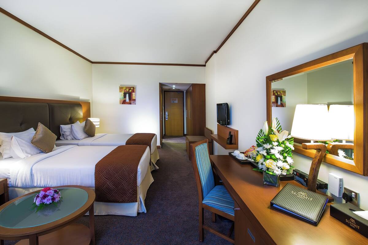 A hotel room with two beds, a desk with a computer, a chair, a small table with a vase of flowers, and a large mirror.