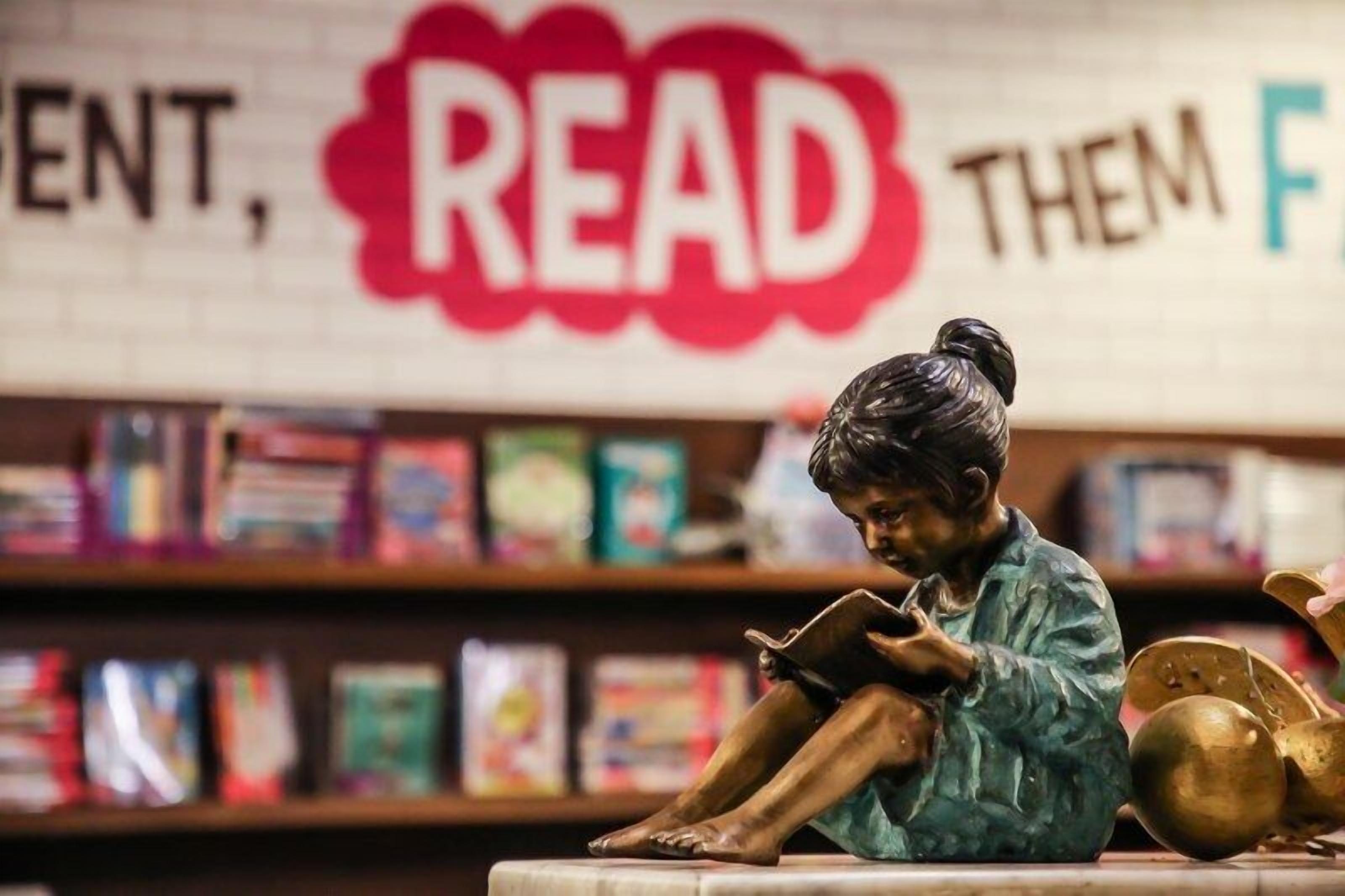 A statue of a person reading in front of a bookshelf with books and a sign that says "READ THEM FIRST".