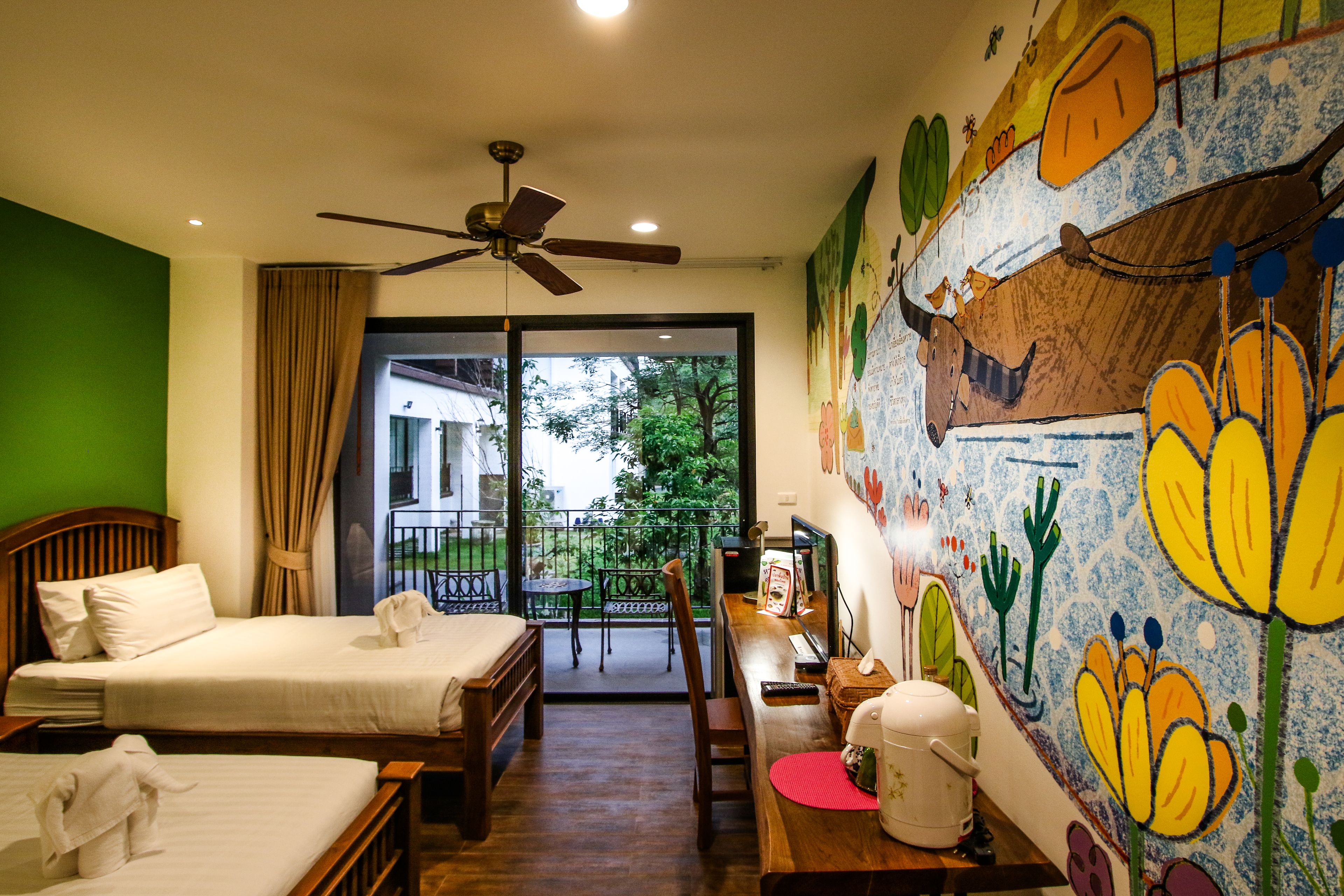 A hotel room with two beds, a ceiling fan, a desk with a chair, a window with a view of greenery, and a colorful mural on the wall.