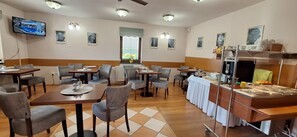 Ein Restaurant mit mehreren Tischen und Stühlen, einer Buffetstation und einem an der Wand befestigten Fernseher.