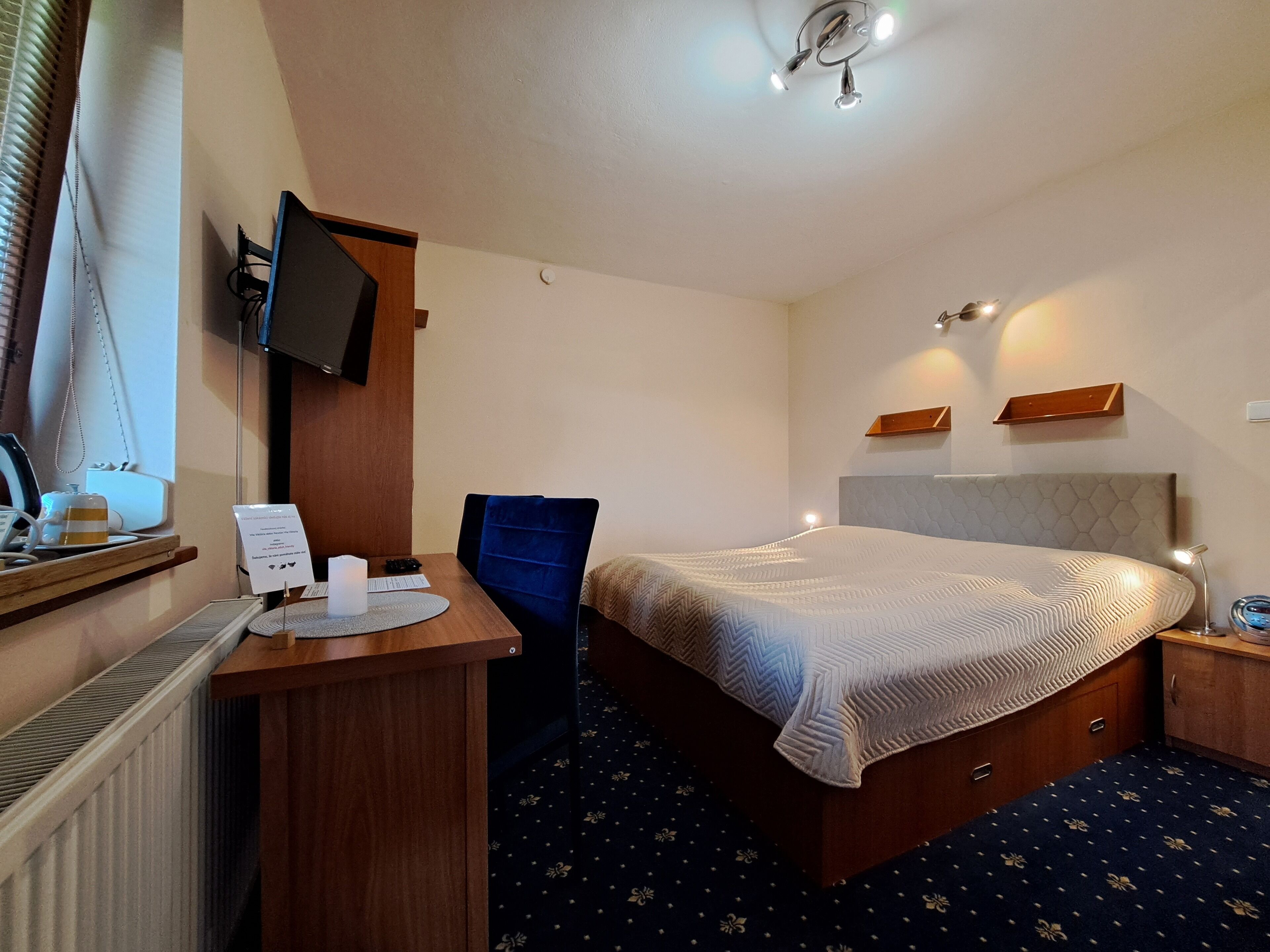 Habitación de hotel con cama, escritorio, silla, televisor y una mesita con una taza y un cartel.