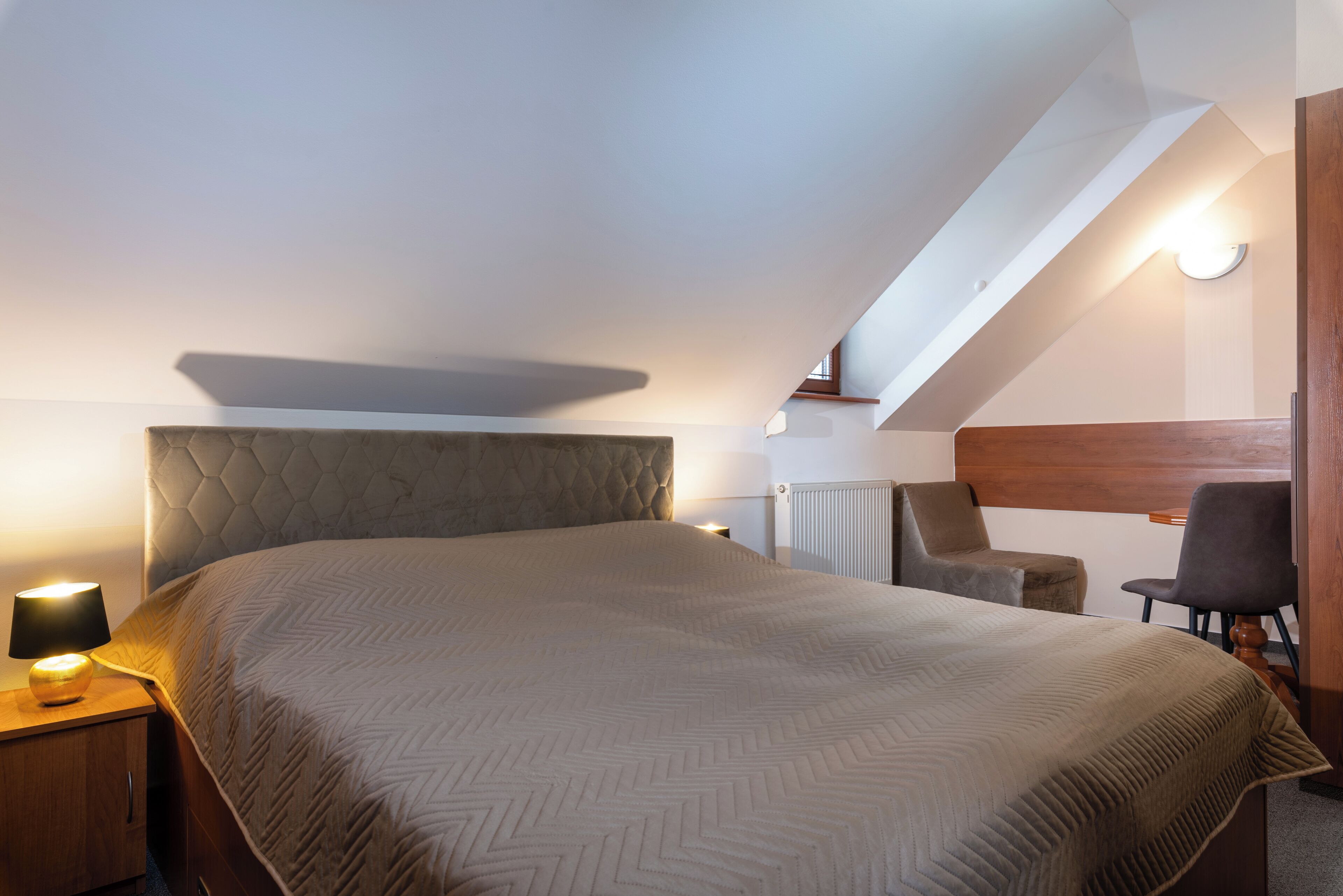A bedroom with a large bed, a nightstand with a lamp, a chair, and a desk.
