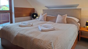 Una cama bien hecha con un cabecero acolchado, una mesita de noche con lámpara y una ventana con persianas.