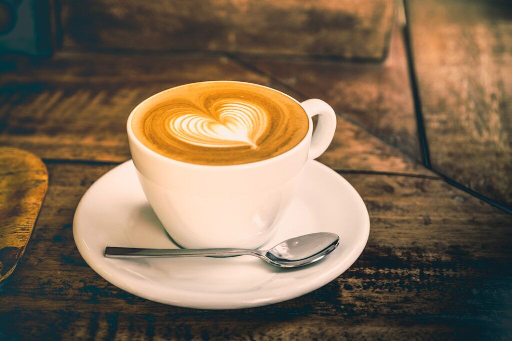 Una taza blanca de café con un dibujo de corazón en la espuma, sobre un platito, acompañada de una cuchara, sobre una superficie de madera.