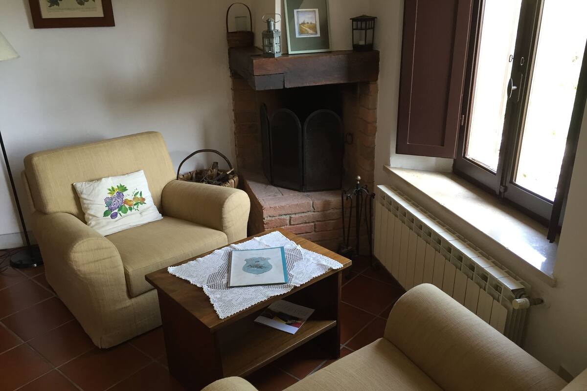 A cozy living room with a beige sofa, a wooden coffee table with a book and a decorative item, a fireplace, and a window with a view.