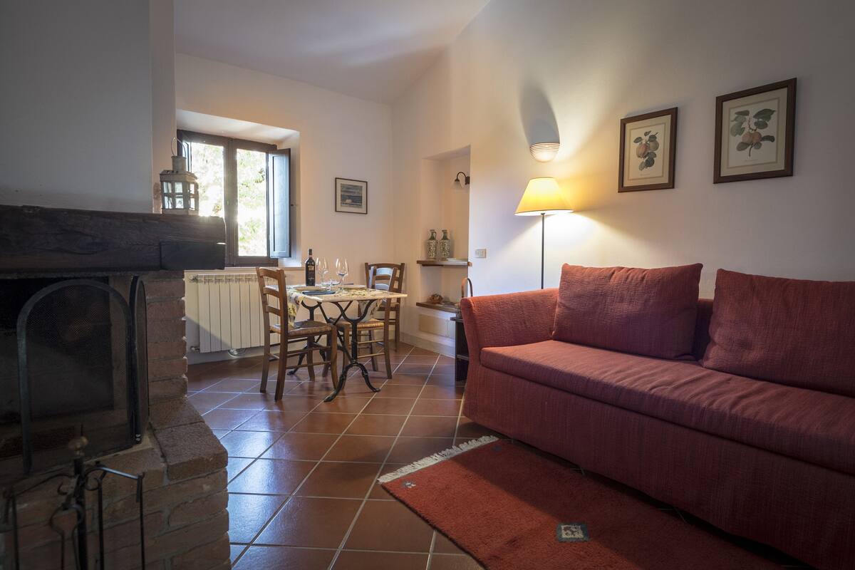 A cozy living room with a fireplace, a red sofa, a dining table with chairs, and a kitchen area in the background.