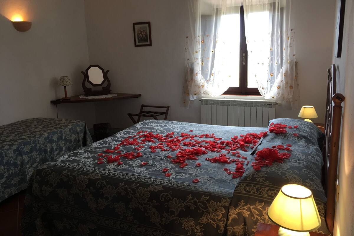A bedroom with two beds, a window with floral curtains, a bedside lamp, and a small table with a lamp.