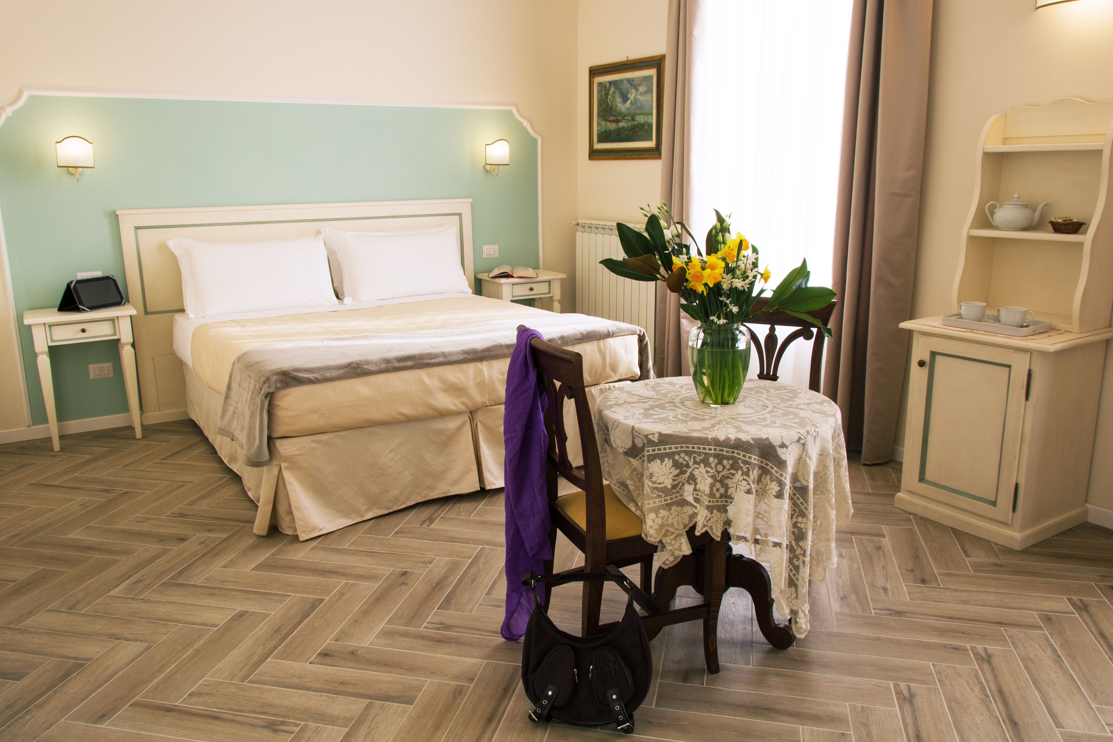 A hotel room with a bed, bedside table, chair, and a small table with a vase of flowers.